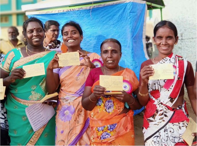 A group of four women stand posing with postcards to the camera. There is a woman and a man also in the background. The four women in the foreground are smiling and are wearing brightly coloured sarees.
