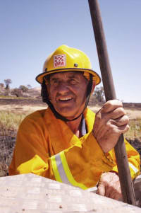 An image of a man wearing firefighting gear. His helmet has the CFA logo and he is looking diagonal to the camera, smiling. He is outdoors, against a field and clear blue sky.