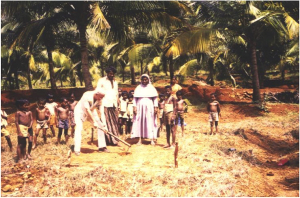 A group of chidren stand in front of palm trees looking towards the camera. Nearby, a nun (Sister Stella) and two other adults stand closer to the camera. One of the adults is digging a well.